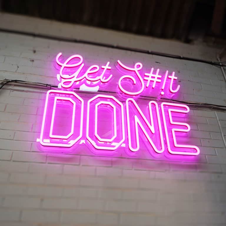 Pink faux neon sign mounted on clear panel. Illuminated sign reads 'Get Shit Done', with custom text options at NeonPlus